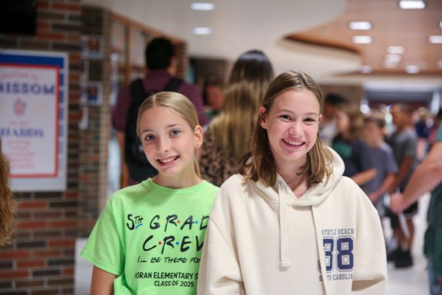 two middle school age girls in hallway of school with other students in the background