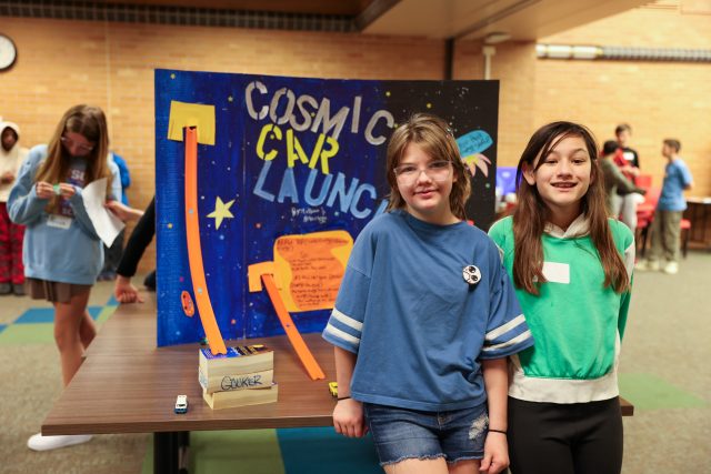 People gathered in a room, engaged in a group activity. Some are seated at tables while others stand around. The atmosphere