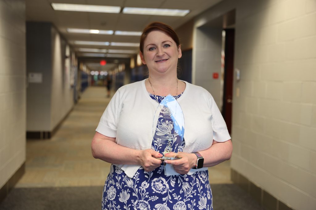 Woman in a blue dress holding an award in front of a hallway.