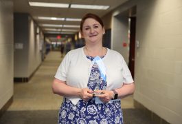 Woman in a blue dress holding an award in front of a hallway.
