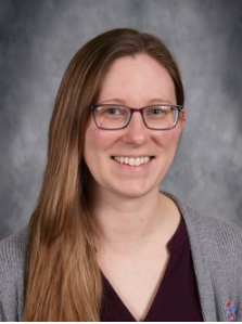 Woman posing for portrait in school setting, smiling slightly, wearing long sleeved black shirt and long brown hair.