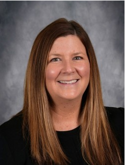 Woman posing for portrait in school setting, smiling slightly, wearing long sleeved black shirt and long brown hair.