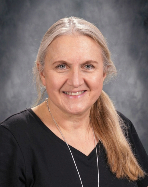 Woman standing in front of a blurred background, smiling at the camera. She has short hair and is wearing a black top.