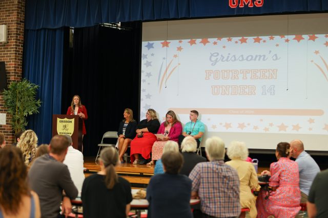 A group of people gathered in a room for an event, standing around a podium and a large screen displaying a presentation.